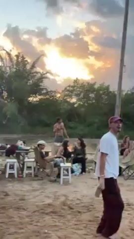 The video shows a group of people gathered outdoors at what appears to be a riverside or beach setting, set against a dramatic sunset sky with bright light piercing through dark clouds. There are trees and other foliage in the background, silhouetted against the sky.  Some individuals are seated at tables and in chairs, engaged in conversation. A shirtless man is seen standing and gesturing with his arms. Another man, wearing a white shirt and a maroon hat, is walking by holding a drink.  Music is playing in the background.