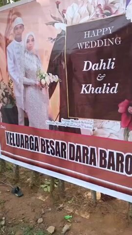 The video shows a series of wedding banners placed outdoors, likely for a celebration. The banners feature a couple in wedding attire, identified as Dahli and Khalid, with "Happy Wedding" and family greetings. The banners are attached to wooden poles or trees, and some are supported by other structures. In the background, there are motorcycles parked, trees, and foliage, suggesting an outdoor setting. There are also glimpses of people, some in modest clothing, possibly attending the event. The ground appears to be dirt and gravel, with some scattered debris. The overall atmosphere suggests a community wedding celebration.