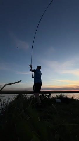 A person is fishing by a body of water at dusk. The person is wearing a blue t-shirt and black shorts and is holding a fishing rod. The sky is blue with some clouds and the sun is setting in the distance, creating a warm glow on the horizon. There is grass and vegetation in the foreground, and the water appears calm. There are also some objects that look like buckets in the background. The overall scene is peaceful and serene.