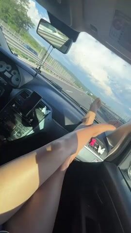 The video shows the interior of a car traveling on a highway. The viewpoint is from the passenger seat looking towards the dashboard and the road ahead. Two bare legs are propped up on the dashboard, obscuring some of the interior of the car. Sunlight streams into the car, casting shadows. A rearview mirror hangs from the windshield, reflecting the road and the sky. The passenger's feet are positioned near the vents of the car's air conditioning system. The driver's hand is visible on the steering wheel. Outside the car, a sunny landscape with green fields and trees can be seen alongside the highway. The sky is blue with scattered clouds. There are decorative butterfly stickers on the dashboard. A smartphone is mounted on the dashboard.