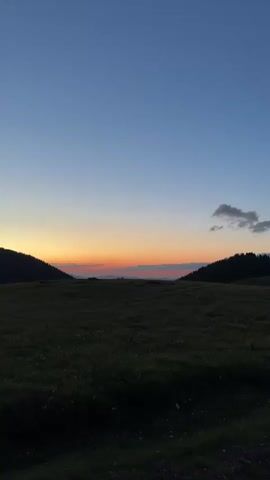 A vertical video shows a landscape at dusk. The sky transitions from a deep blue at the top to a soft orange and yellow near the horizon. Rolling hills, covered in dark grass, frame the sunset. The foreground is a field of grass, dark and shadowed. The overall impression is one of natural beauty and tranquility.