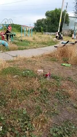 The video shows a playground in Belovodskoe, Kyrgyzstan. Three young people are interacting with playground equipment. Two girls, one in a yellow outfit and the other in an orange shirt and black pants, are on a seesaw. A third person, also in an orange shirt, is near the seesaw. The playground features two slides and a tire swing. The setting appears to be an outdoor park with trees and dry grass. The weather seems overcast. The overall atmosphere is casual and playful, with the individuals appearing to be enjoying themselves. There are no visible signs or text in the video.