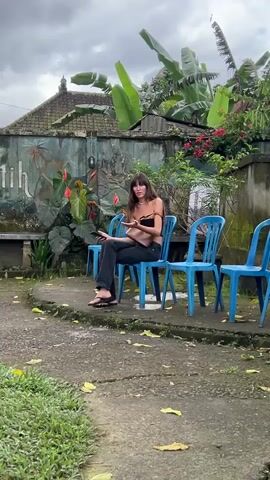 Here's a description of the video:

A woman is sitting on a light blue plastic chair in an outdoor setting. She is holding a smartphone in her hand and appears to be using it. She is wearing a black strapless top, dark jeans, and sandals. A tan purse is slung over her shoulder. Several similar empty chairs are positioned around her.

The environment includes a low building with a painted mural on its wall, tropical plants, and a stone or concrete ground covered with some leaves and patches of grass. The sky is overcast.