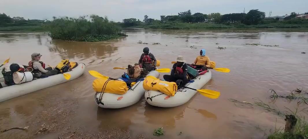 The video shows a group of people preparing to embark on a river rafting trip. There are three inflatable rafts near the muddy shore of a river. The rafts are loaded with gear, including yellow dry bags, backpacks, and paddles. People are sitting in the rafts, wearing life vests, and appear to be getting ready to push off. A man in a red life vest walks along the shore towards the rafts. The water in the river is brown and murky. The background shows a green riverbank with trees. The sky is overcast.