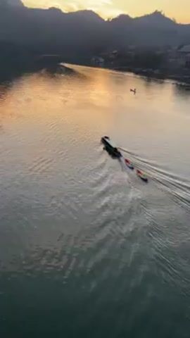 The video shows a boat moving along a wide river with two brightly colored kayaks tethered behind it. The boat leaves a wake on the water's surface. The river reflects the golden light of the setting sun. The opposite bank of the river is visible, with trees and a few buildings nestled on the shoreline. There is another small boat with a person standing on it, further up the river. The camera appears to be positioned above the river, possibly on a bridge or elevated platform.