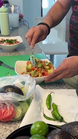 A person is preparing food on a kitchen counter. The person, wearing a black t-shirt with red car designs and a braided black bracelet, is mixing a salad in a white bowl with a spoon. The salad appears to contain avocado and tomato. Nearby on the counter, there is a plastic bag filled with limes and tomatoes, and a paper towel with green chili peppers on it. There is also a black bowl filled with avocados and limes. In the background, a green water bottle and a glass of milk are visible. Another salad bowl is also visible.