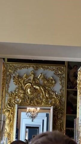A video displays a grand interior space, possibly a museum or historical building. The first frame shows a gilded ornate relief above a doorway, with a painting visible through the opening. The subsequent frames focus on a large, elaborate chandelier with multiple light bulbs, hanging from the ceiling. Below the chandelier, two framed paintings hang on a light-colored wall. The paintings appear to be portraits of men in historical attire. The overall impression is one of opulence and historical significance.