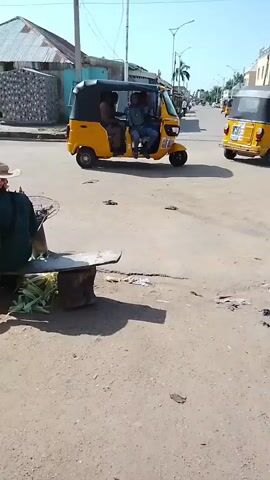 The video shows a street scene with two yellow auto-rickshaws. One auto-rickshaw is in the foreground, and another is in the background. Both are moving away from the camera. There are people in the auto-rickshaws. The street appears to be unpaved, and there are some buildings on the side of the road. The weather is sunny. There is also what appears to be a person sitting on a stool in the foreground, near some produce.