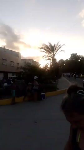 The video shows a street scene at dusk. The sky is filled with clouds, and the sun is setting, casting a warm glow. A palm tree stands prominently in the foreground, silhouetted against the sky. On the left side of the street, there are buildings and several people are visible, some sitting and others standing. A car is parked on the right side of the street. The road is paved and has a yellow curb. The overall atmosphere is serene and calm.