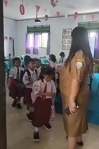 A teacher in a tan uniform is in a classroom with several young students in school uniforms. The teacher is standing on the right side of the frame, facing a line of children. The children are in a line, facing the teacher. The teacher is holding a phone in her right hand. One child in the front of the line extends their hand towards the teacher. The classroom has desks, chairs, and decorations on the walls.