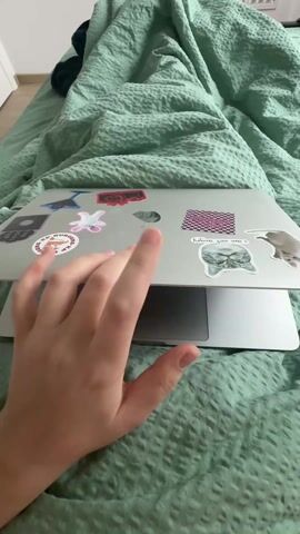 A hand opens a silver MacBook laptop on a bed with a light green patterned duvet. The laptop is covered in stickers. The hand opens the laptop so that the screen is halfway up.