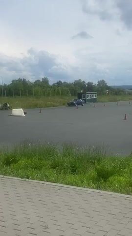 A blue hatchback car is seen driving on a paved surface, possibly a track or an empty lot. The car is blue and appears to be a later model hatchback. There are several orange traffic cones placed around the area, suggesting a driving course or training session. The car is performing a maneuver, possibly a drift or a sharp turn, as indicated by the circular tire marks on the ground. In the background, there are trees, a fence, and a cloudy sky. The foreground shows green grass and a paved walkway. Another car, black, is seen briefly at the end of the clip, also on the paved surface.