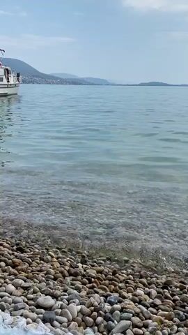 A shot of a pebble beach and calm water. A boat is visible on the left side of the frame, floating on the water. The sky is clear and blue with a few clouds. The water is clear enough to see the pebbles on the bottom of the lake or sea.