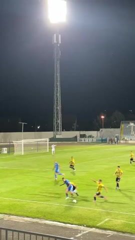 A soccer game is being played at night under stadium lights. The field is green grass, and there are players in blue and yellow uniforms. A tall light tower is visible in the background. The players are engaged in a play, with the ball on the ground.