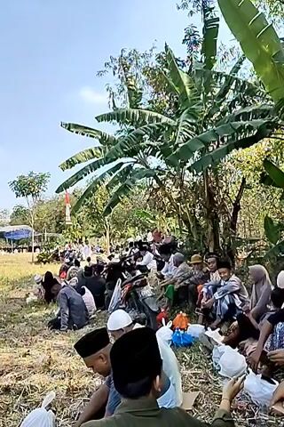 A group of people, appearing to be predominantly male and of varying ages, are seated on the ground in an outdoor, grassy area. They are gathered under trees, with lush green foliage visible, including large banana leaves. Some individuals are wearing traditional Islamic attire, such as kufi hats. They seem to be engaged in a communal activity, with some interacting with each other and others appearing to be listening or observing something off-camera. Food items and bags are scattered around them. In the background, there are tents and structures, and a flagpole with a red and white flag. The sky is clear and blue, indicating a sunny day. The overall atmosphere appears to be one of a gathering or event.