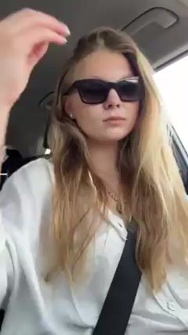 A young, light-skinned woman with long blonde hair sits in the driver's seat of a car, wearing dark sunglasses, a white button-down shirt, and a seatbelt. She runs her fingers through her hair, checks her appearance, and puckers her lips slightly. The car's interior is visible, including the dashboard and a window showing an overcast sky.
