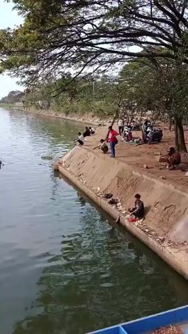 A medium-shot video captures a canal with several people fishing along its edge. The water is calm and green, reflecting the trees and sky. The bank of the canal is lined with concrete, and several individuals are seated, casting lines into the water. Some are young, while others appear to be adults. Further back, a few people are gathered near motorcycles and bags, suggesting a leisurely outing. A bridge can be seen in the distance, spanning the canal. The overall atmosphere is peaceful, with a sense of community as people engage in a shared activity.
