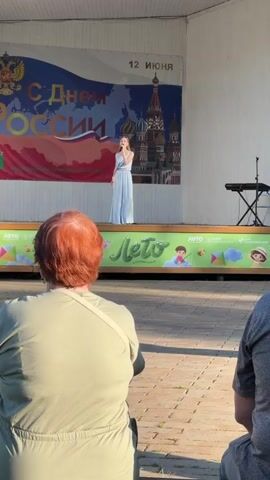 A video shows a wide outdoor shot of a woman in a light blue jumpsuit singing on a stage with a microphone. Her light brown hair is tied back in a ponytail. The stage backdrop is a banner that says "С Днем России" (Happy Russia Day) and "12 ИЮНЯ" (June 12), with an image of a map of Russia and St. Basil's Cathedral. Below the stage, a decorative banner says "Лето" (Summer) in green with drawings of children. In the foreground, facing away from the camera, is a woman with red hair wearing a light green shirt and a white cross-body bag. To her right, the shoulder of another person wearing a gray shirt is visible. The ground in front of the stage is paved with light brown bricks. The video is still and does not show any movement.