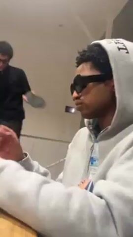 The video depicts three young men in what appears to be an indoor setting. In the foreground, a man wearing a grey hoodie and dark sunglasses sits at a wooden table looking at his cell phone. In the background, a man dressed in black with sunglasses dances and throws a black object in the air. As the camera angle shifts, a third person, wearing a pink cap and a black t-shirt with Arabic writing, is seen standing by the table. The overall mood appears lively and playful.