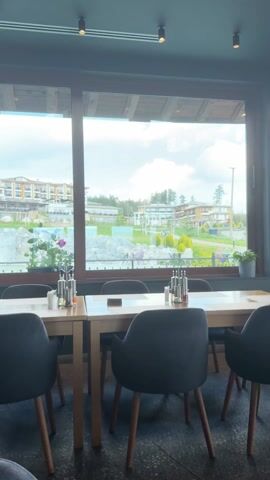 The video shows an interior view from a restaurant with a large window overlooking an outdoor scene. In the foreground, there are several dark-colored chairs arranged around wooden tables set for dining. Each table is equipped with salt and pepper shakers, and a potted plant sits on the windowsill. The large window offers a view of a well-manicured landscape featuring a decorative waterfall, large rocks, and a grassy area. Several buildings, possibly hotel or resort structures, are visible in the background. The sky is blue with scattered clouds.