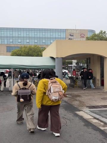 The video shows a group of people, mostly young, walking into a building through a covered entrance. Several people are wearing backpacks. The building has a sign in what appears to be Chinese characters, with an English translation below it. The weather looks overcast. Some red crowd control barriers are visible on the left, and scooters are parked in the background. People are walking in various directions.