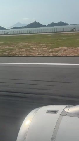 The video shows an aerial view of a runway with an airplane engine in the foreground. The plane is on the runway and appears to be taking off or landing. The runway is asphalt and has white markings. Beyond the runway is a grassy area with a white fence. In the background, there are mountains and the sea. The sky is clear and blue. The sun is shining and reflecting off the airplane engine. There are no people visible in the video.
