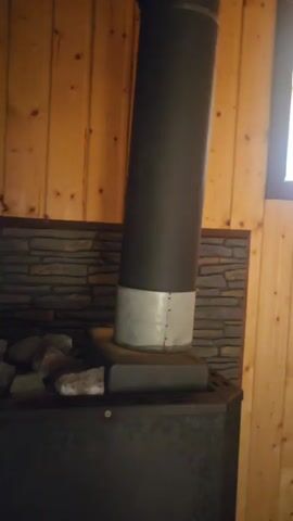 The video shows the interior of a wooden room, possibly a sauna. The walls and ceiling are paneled with light-colored wood. In the first frame, a dark metal stovepipe emerges from a dark stove. There is a stone-like pattern on the wall behind the stovepipe, and a window with bright light coming through it is visible on the right. The subsequent frames show benches made of wooden slats. In one shot, a large metal basin is visible under a bench. In another shot, a wooden ladle rests on a bench. The room appears to be empty of people.