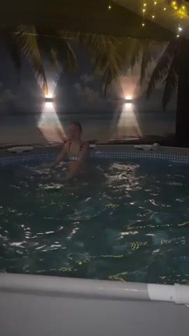 A young girl in a bikini stands in a round, above-ground pool at night. The pool is filled with clear water. Behind her, a backdrop displays a beach scene with palm trees under a twilight sky. Two wall-mounted lights cast beams onto the backdrop, enhancing the tropical illusion. String lights are strung above the scene. The girl looks towards the camera, gently moving her arms in the water. The pool has a white plastic rim.