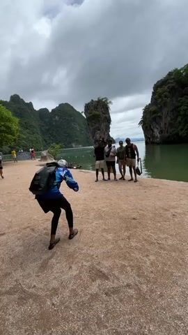 A group of five individuals stands on a sandy shore, with a person in the foreground facing them. The group of five are standing close together, some holding bags and others with hands in pockets. The person in the foreground, wearing a backpack and a blue jacket, is facing away from the camera and appears to be gesturing towards the group. Behind the individuals, a large rock formation juts out of the emerald green water, flanked by lush, green, mountainous islands. The sky above is overcast with dramatic clouds. Another person is visible in the background on the left, walking towards the shore.
