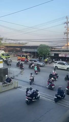 The video shows a busy street scene with a lot of motorcycle traffic. There are also a few cars and a bus. The motorcycles are moving in different directions, and some of them are carrying multiple passengers. There are also a few pedestrians walking on the side of the road. The overall impression is one of a chaotic but functional urban environment. The scene appears to be taking place in a warm climate, as people are dressed in light clothing. The sky is clear, and the sun is shining. The buildings in the background appear to be a mix of commercial and residential structures. There are also a lot of power lines and utility poles, suggesting a developed area. The video seems to be shot from a slightly elevated position, giving a good overview of the traffic flow.