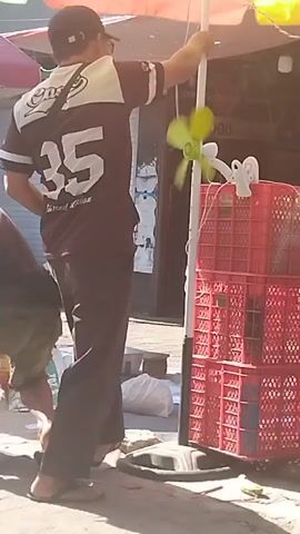 The video shows two men in an outdoor market setting. One man, wearing a dark t-shirt with white stripes and the number 25 on the back, a black baseball cap, and sunglasses, is standing and adjusting a white umbrella pole that has a small green fan attached to it. He is wearing dark pants and flip-flops. The other man, on the left side of the frame, is bent over and appears to be interacting with large bags of goods. The environment includes market stalls, red plastic crates, and various bags and boxes on the ground. The lighting suggests it is daytime.