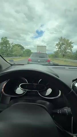 The video shows two separate shots inside cars. The first shot is from the driver's perspective, looking out the windshield at a gray Peugeot driving in front of the car on a highway. Trees are visible on either side of the road, and the sky is cloudy with patches of blue. The second shot is from the passenger seat, looking up at a young woman wearing a red long-sleeved shirt and jeans. She is wearing a seatbelt, gold hoop earrings, and a necklace. She has light brown hair and is looking towards the camera. She raises her hand to touch her head. The car interior is red and black.