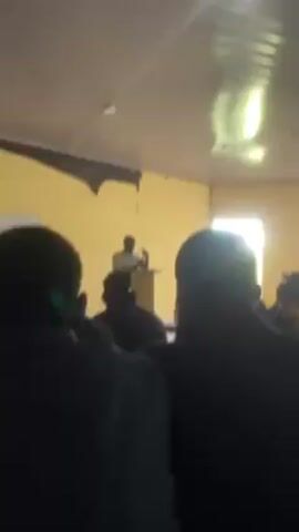 The video shows a person standing behind a podium addressing an audience in a classroom setting. The classroom has yellow walls and barred windows. The audience members are facing the speaker, with their backs to the camera. There are ceiling fans in the room. The speaker is gesturing with their hands while speaking.