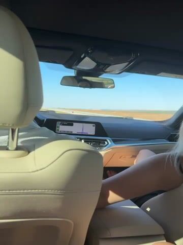 The video shows the interior of a car from a rear passenger's perspective. The dashboard and front windshield are visible, displaying a navigation system. A person with blonde hair is in the driver's seat, facing away from the camera. Their arm is resting on the center console. Another person's legs are visible in the foreground, suggesting they are in the rear passenger seat. The car appears to be driving on a road with a flat, open landscape visible outside. The sky is blue and clear. The lighting suggests it is daytime.