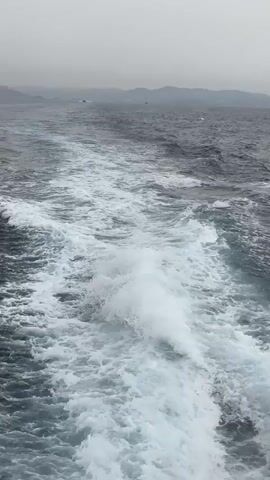 A vertical, medium shot captures the turbulent wake of a boat moving through the ocean on an overcast day. The camera is positioned at the stern of the vessel, looking backward. The water is a deep, dark blue-gray, churned into a frothy white trail by the boat's passage. Waves are visible in the foreground, breaking and swirling with foam. In the distance, the horizon is marked by a hazy, gray sky meeting the darker expanse of the sea. The outline of land, possibly islands or a coastline, is faintly visible in the far distance, with a few small boats or buoys scattered on the water. The overall atmosphere is somber and slightly choppy, suggesting movement and a moderate sea state.