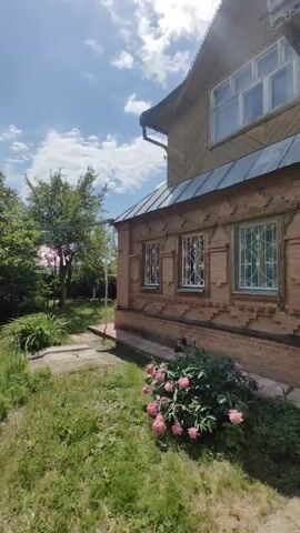 The video shows a partially shaded view of a house with a brick lower story and wooden upper story. The house has several windows with white frames and decorative metal grates. In front of the house, there is a bush with pink flowers, and a lawn with patches of dirt and gravel. Trees are visible in the background, and the sky is blue with white clouds. The sun appears to be shining.