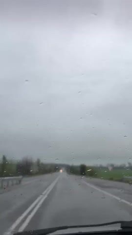 The video shows a first-person perspective from inside a car driving on a wet asphalt road in Cherganovo, Bulgaria. Raindrops are clearly visible on the car's windshield, indicating it is raining. The sky is overcast and gray. The road features white double lines down the center. In the distance, the headlights of another vehicle can be seen. Trees and fields line both sides of the road, appearing somewhat blurred due to the rain and distance. Upbeat music is playing in the background. No people are visible, and there is no discernible text. The scene depicts a drive during rainy, daytime weather.