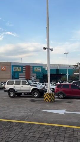 The video displays an outdoor scene in what appears to be a commercial or urban area in Guayana City, Venezuela, during daylight hours under a blue sky with scattered clouds.

The initial shot shows a parking lot with several parked cars, including a silver SUV and a red hatchback, on concrete asphalt marked with a white arrow pointing left and a yellow striped curb. A tall white pole with speakers stands prominently in the foreground. In the background, a building complex with a blue and green roof is visible, along with a white building featuring the text "MYKONOS."

As the video pans and zooms, a wider view of the parking lot reveals more vehicles, including a black car driving away. The "MYKONOS" building, possibly a restaurant or shop, is more clearly seen amidst palm trees.

The final part of the video focuses on a distant, tall, cylindrical building made of glass panels. This building is identified by a rooftop sign that reads "Hotel Plaza Meru." A distinctive concrete structure resembling steps or terraces is attached to its side. Palm trees frame the hotel, reinforcing the warm climate. No people are clearly visible in the video, emphasizing the architectural and environmental elements of the scene.