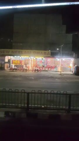 The video shows a night-time view of a restaurant called "NEW NEELAM DHABA" located on the side of a road. The restaurant is lit up with yellow and white lights. There are tables and chairs set up outside the restaurant. Signs indicate "VEG THALI" and "FAST FOOD". A tall, grey building is visible behind the restaurant. A metal fence runs across the foreground of the video. The image is slightly blurry.