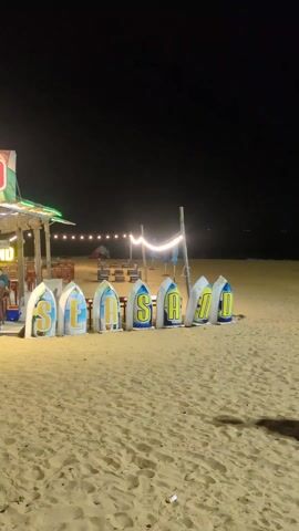 Here is a description of the video:

The video shows a beach at night. In the foreground, there are large surfboard-shaped signs spelling out "SAUDI". Further back, there are scattered groups of people walking and standing. In the distance, across the water, there are lights visible on the horizon. A person in an orange jacket walks from left to right, and another person in a black jacket and jeans walks from right to left. The sand is light in color, and there are several shadows visible. The sky is dark and clear.
