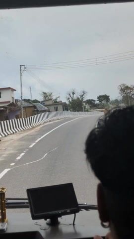 From within a vehicle, a view of a road curving slightly to the right unfolds. A two-story building labeled "EMC HOSPITAL" is visible on the left. The roadside is marked by a black and white striped barrier. Overhead, a network of power lines stretches across a cloudy sky. In the foreground, atop the dashboard, sits a small, decorative golden shrine and a black monitor. The back of a person's head is visible on the right side of the frame.