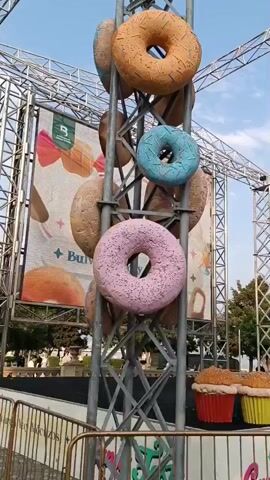 The media file captures an outdoor scene, likely at an event or amusement area. Dominating the foreground is a tall, metal truss structure adorned with large, oversized donuts in various colors and flavors: orange, blue, and pink. Behind this confectionery display, a large screen or banner features stylized images of donuts and ice cream, with text that appears to include "B" and "Burg".

Several people are visible in the mid-ground. Two young girls are walking and eating. One in a white t-shirt and jeans is holding what looks like a small cake or pastry. Another girl in a pink polo shirt is also holding a dessert. To their right, a woman wearing a hijab and a black and white patterned top is seen holding a piece of food.

Further back, beyond a yellow metal barrier with signage that includes "Candy Fest", are more giant props resembling cupcakes with yellow and red bases and brown tops. Behind these, a line of trees borders a body of water, suggesting a seaside or lakeside location. In the distance, several individuals are walking along a pathway, and a man in a maroon shirt and blue jeans is walking to the right.

The sky is a clear blue with a few scattered clouds, indicating fair weather. The lighting suggests it is late afternoon or early evening. The overall atmosphere appears festive and family-friendly, with the giant food props and the presence of people enjoying treats contributing to a cheerful ambiance. The setting seems to be a public recreational area or a temporary event space.