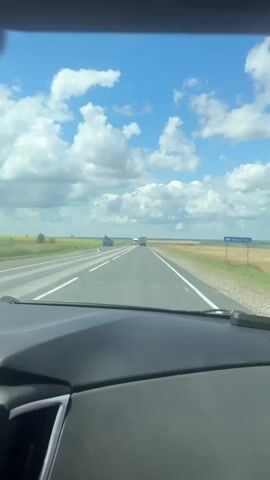The video is taken from the perspective of someone driving a car, looking out through the windshield. The road ahead is a multi-lane highway with a clear blue sky and fluffy white clouds above. The landscape on either side of the road appears to be flat farmland, with yellow and green fields stretching to the horizon. Several trucks and cars are visible on the road ahead. One large white truck is in the left lane, and several other vehicles are further down the highway. A traffic sign is visible on the right side of the road. The dashboard of the car is visible in the foreground. The lighting suggests it is daytime.