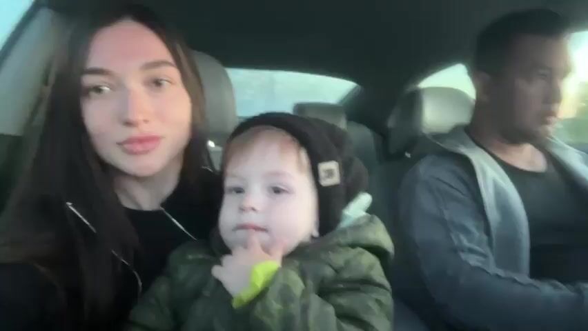 Inside a car in Пермяки, Russia, a young family is captured in a close-up shot. A woman with long dark hair, wearing a black top, is seated on the left, looking directly at the camera with a gentle smile. She then turns to interact with the child beside her.

In the center, a young child, likely a toddler, is seated between the woman and the man. The child is wearing a black knitted beanie with a distinctive white letter 'R' on it, and a camouflage-patterned jacket with green accents. Initially, the child looks towards the camera, then turns their attention to the woman, pointing at her face. The child also brings a finger to their mouth at one point.

On the right, a man with dark hair, dressed in a grey jacket over a black shirt, is in the driver's seat. He appears focused forward, possibly on the road or a task, and is seen unwrapping what looks like a snack or item.

The setting is the interior of a car, suggesting they are either traveling or parked. The light visible through the car windows indicates it is daytime, possibly late afternoon or early evening, with clear conditions outside. The overall mood appears calm and domestic.