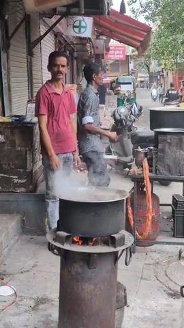 A man in a red shirt stands next to a large pot of liquid boiling over an open flame on a metal stove outdoors. Another man in a patterned shirt stands behind him, with his hand to his ear. In the background, there are shops, a motorcycle, and a propane tank. The scene appears to be a street vendor preparing food or drink.