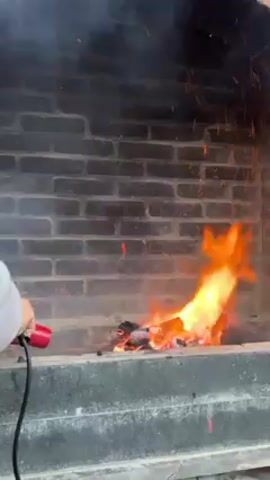 A fire burns in a brick fireplace. Someone is using a red tool to fan the flames, which are orange and yellow. There is smoke and sparks rising from the fire. The fireplace is made of gray bricks and a gray concrete base. The tool has a black cord.