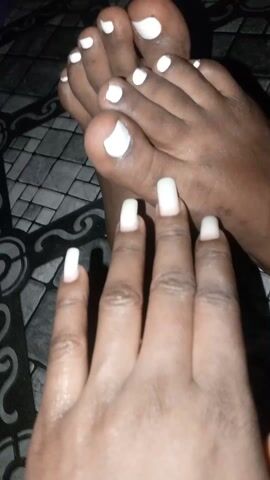 A pair of dark-skinned feet with white nail polish on the toenails is positioned near a hand with similarly colored, long nails. The hand moves slightly, revealing more of the palm. The background is a patterned surface.