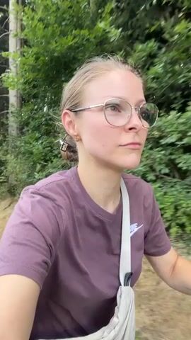 A woman with blonde hair pulled back is wearing glasses and a purple t-shirt. She is on a bicycle, and the video shows her face close up before panning out to reveal a scenic view of rolling green hills, a field, a road, and a treeline under a bright blue sky with fluffy white clouds. The woman has a crossbody bag over her shoulder.