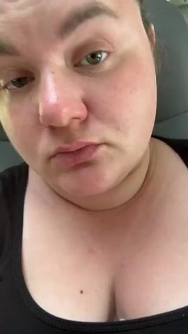 The video shows a close-up of a person's face and upper chest. The person has light skin, green eyes, and brown hair pulled back. They are wearing a black tank top. The background appears to be the interior of a vehicle, with a grey seat visible. The lighting is natural and appears to be daylight. The person's head moves slightly throughout the video.