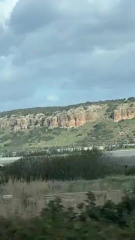 The video shows a landscape with a cliff visible in the background. The cliff is composed of rock formations and is covered in green vegetation on top. The video is taken from a moving vehicle, as seen by the passing trees and plants in the foreground. The sky is cloudy with patches of blue. A utility pole is visible in front of the cliff, suggesting the presence of infrastructure.